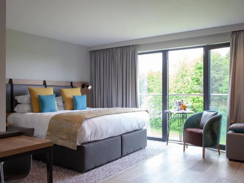 Modern hotel bedroom with a double bed, colourful cushions, armchair and large glass doors overlooking garden greenery