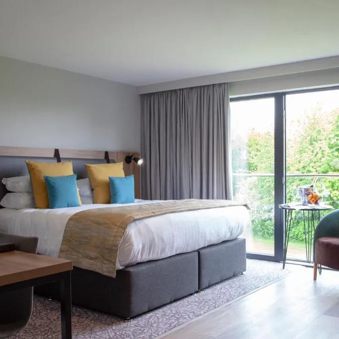 Modern hotel bedroom with a double bed, colourful cushions, armchair and large glass doors overlooking garden greenery