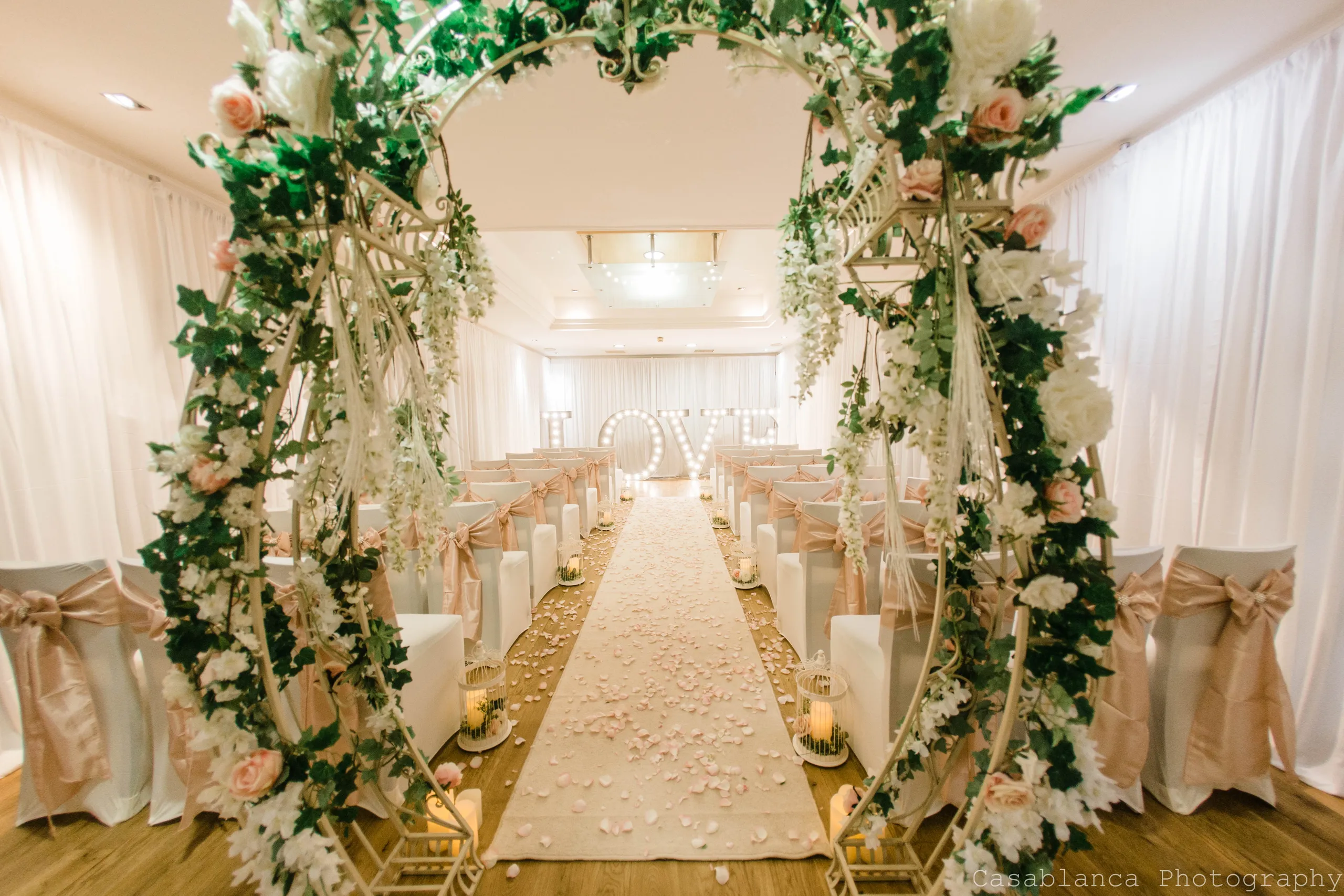 Elegant wedding ceremony room with a flower-covered arch, petal-strewn aisle and illuminated LOVE letters.