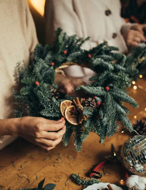 Two people making festive evergreen wreaths with dried oranges, cinnamon and fairy lights at a wooden table.