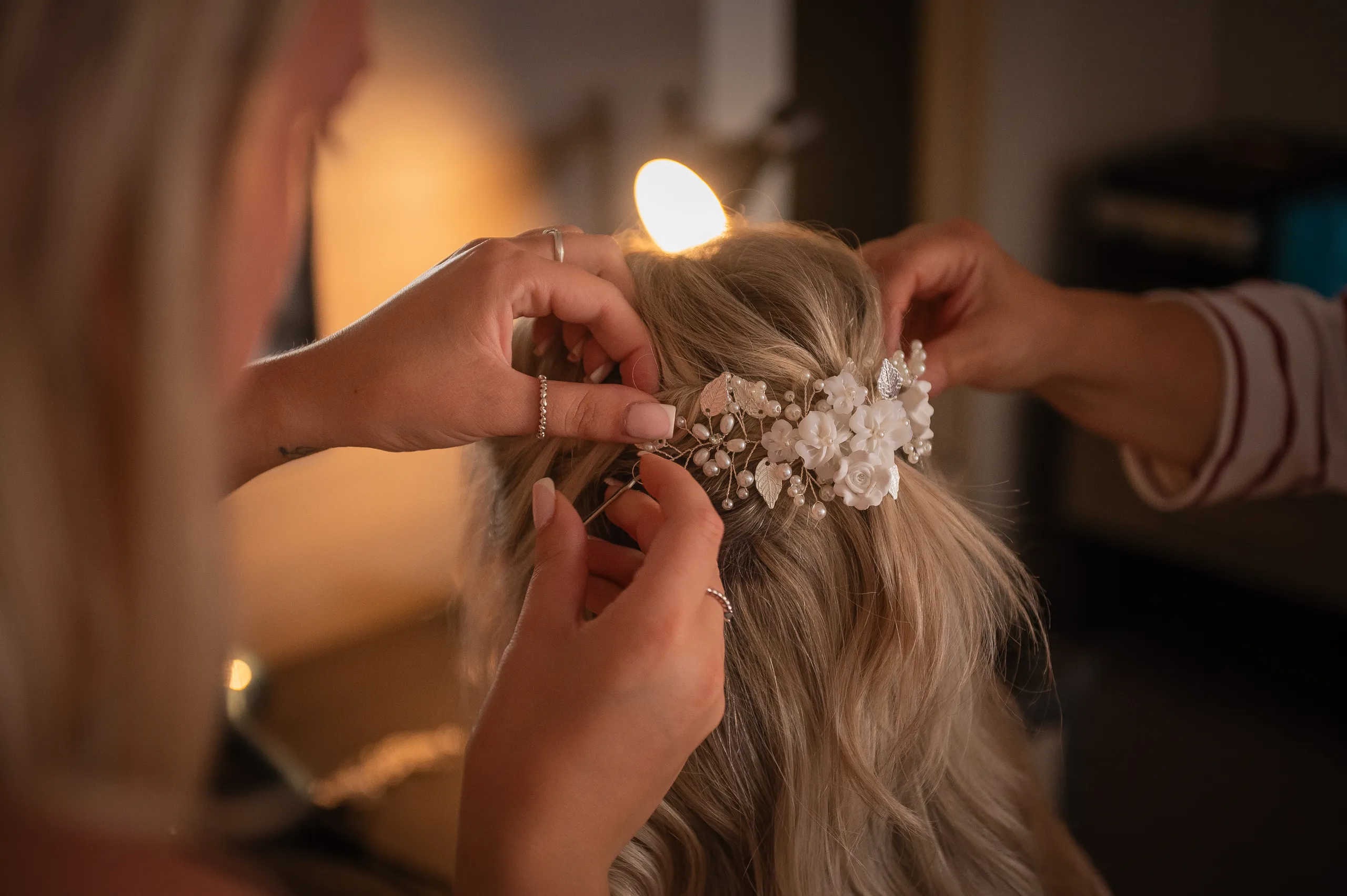 Hands fastening a delicate white floral and pearl hairpiece into a bride's blonde updo