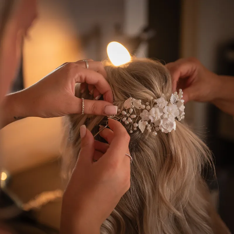 Hands fastening a delicate white floral and pearl hairpiece into a bride's blonde updo
