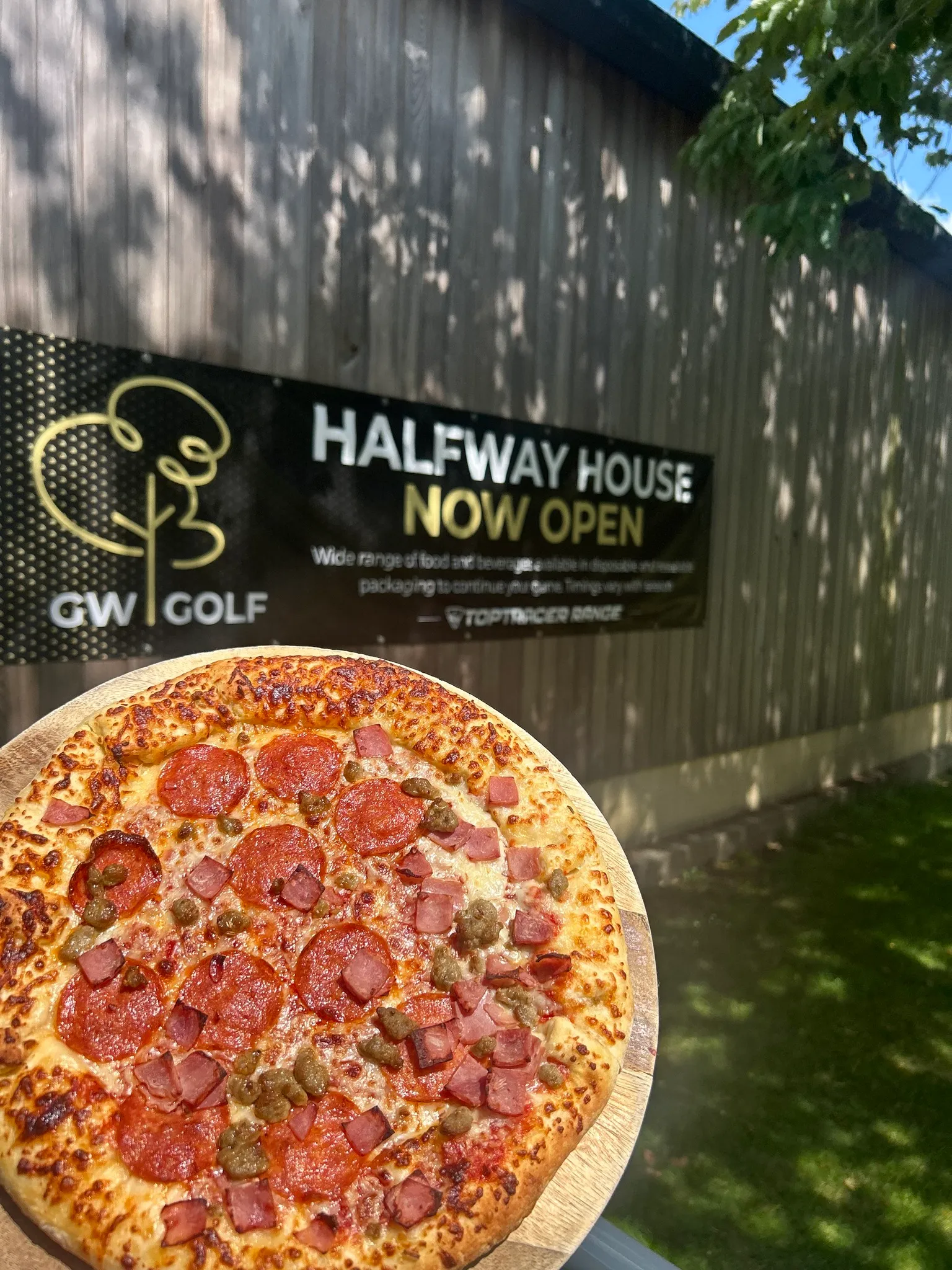 Pepperoni, ham and sausage pizza held in front of a GW Golf Halfway House sign.