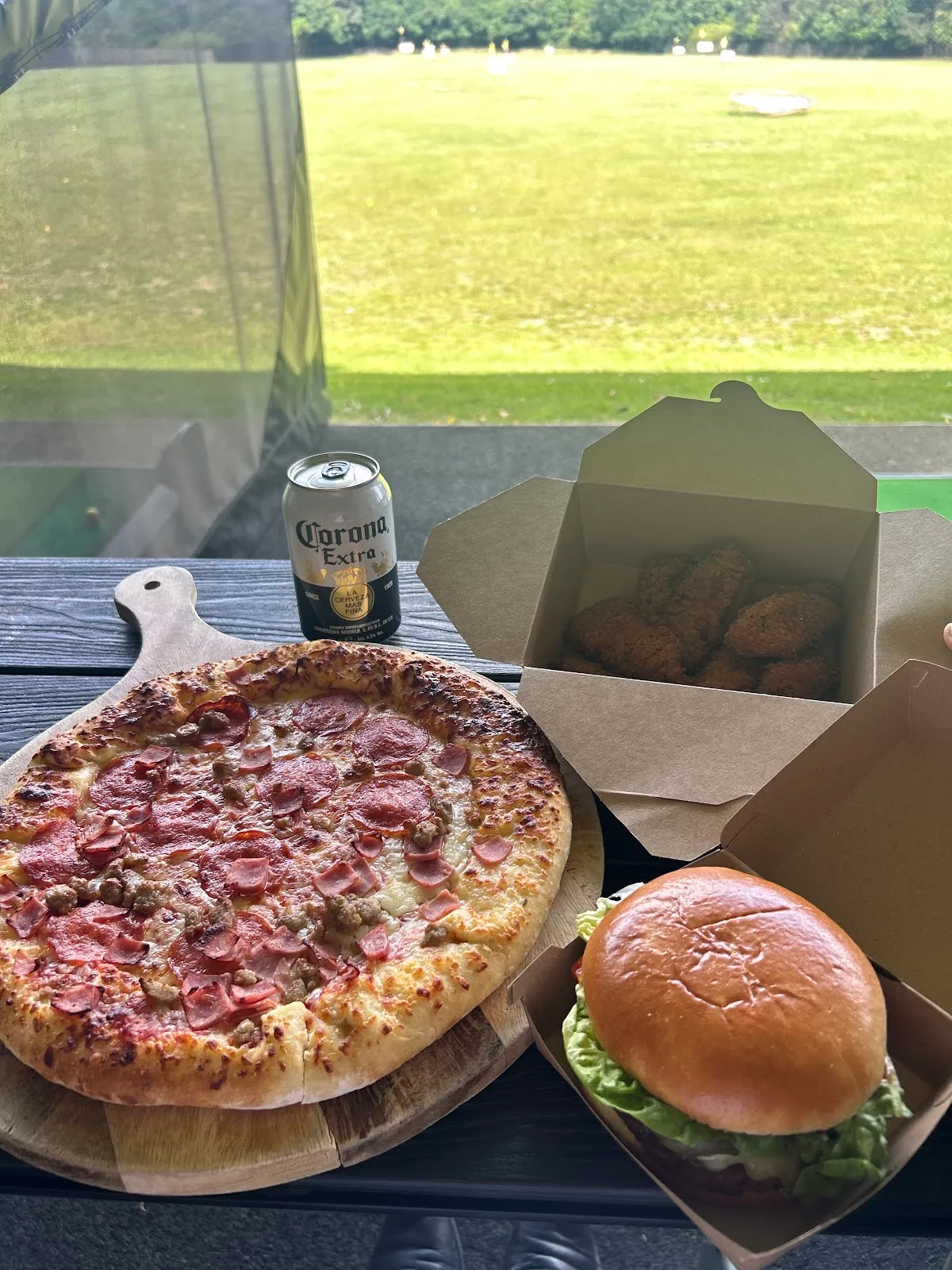 Outdoor table spread with pepperoni pizza, burger, fried chicken and a can of Corona Extra beer.
