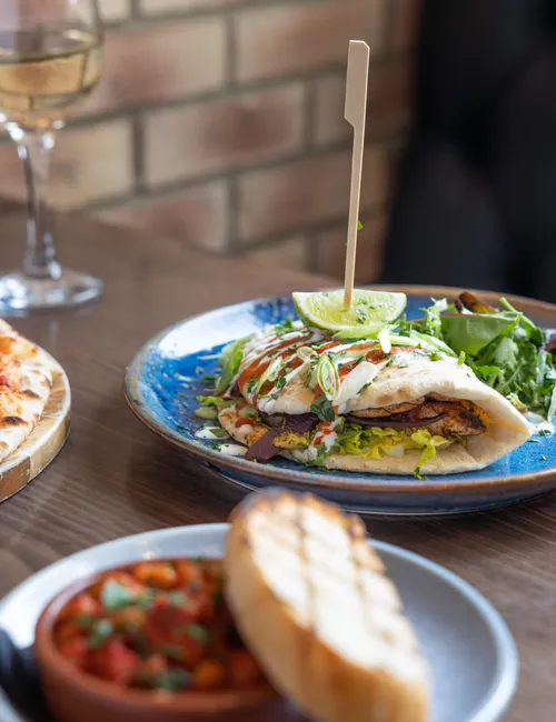 Grilled chicken flatbread with salad, lime and sauces on a blue plate beside pizza and a glass of white wine.