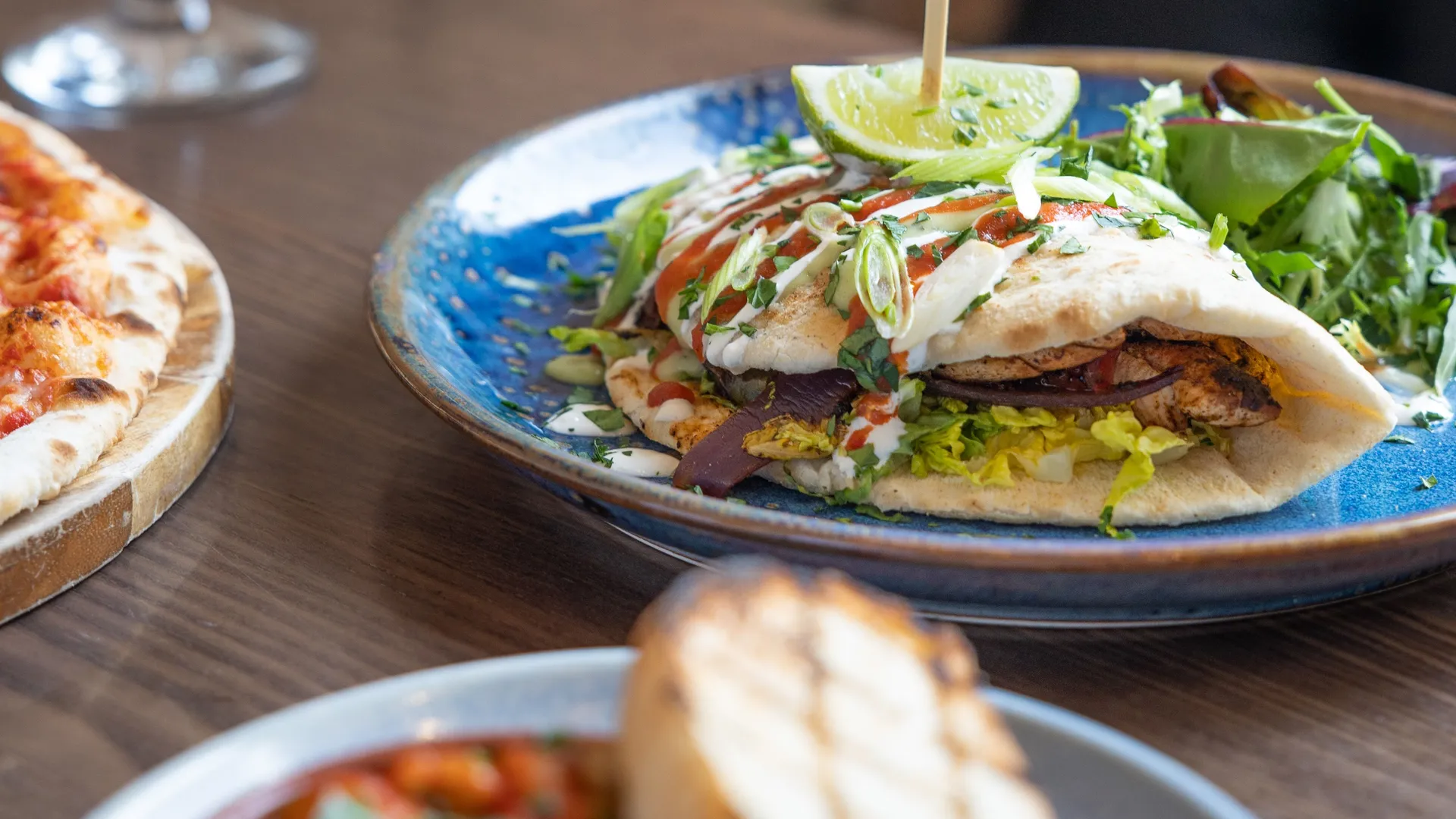 Grilled chicken flatbread with salad, lime and sauces on a blue plate beside pizza and a glass of white wine.