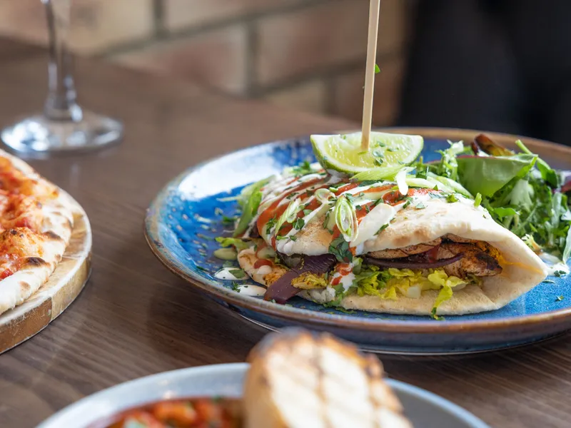Grilled chicken flatbread with salad, lime and sauces on a blue plate beside pizza and a glass of white wine.