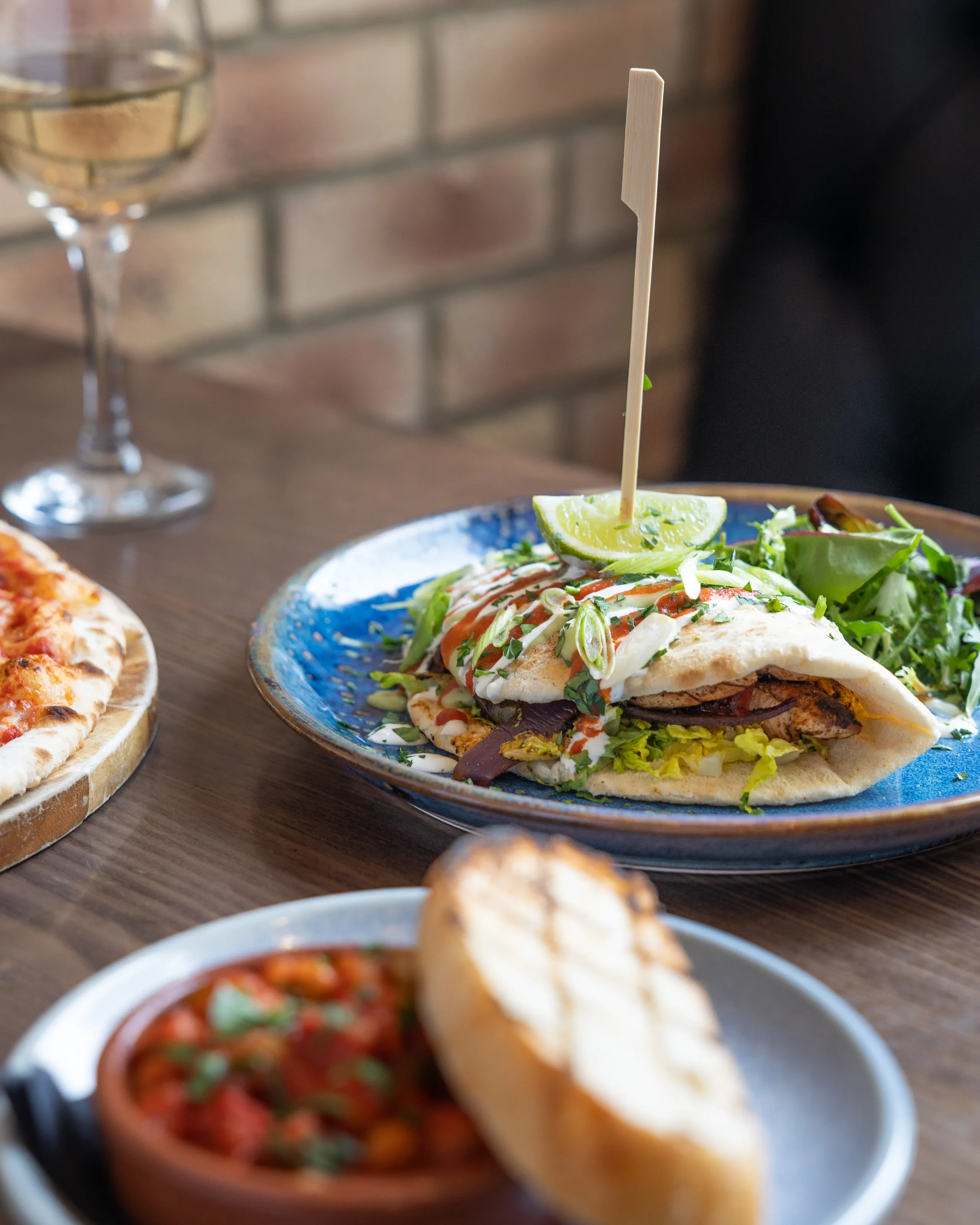Grilled chicken flatbread with salad, lime and sauces on a blue plate beside pizza and a glass of white wine.