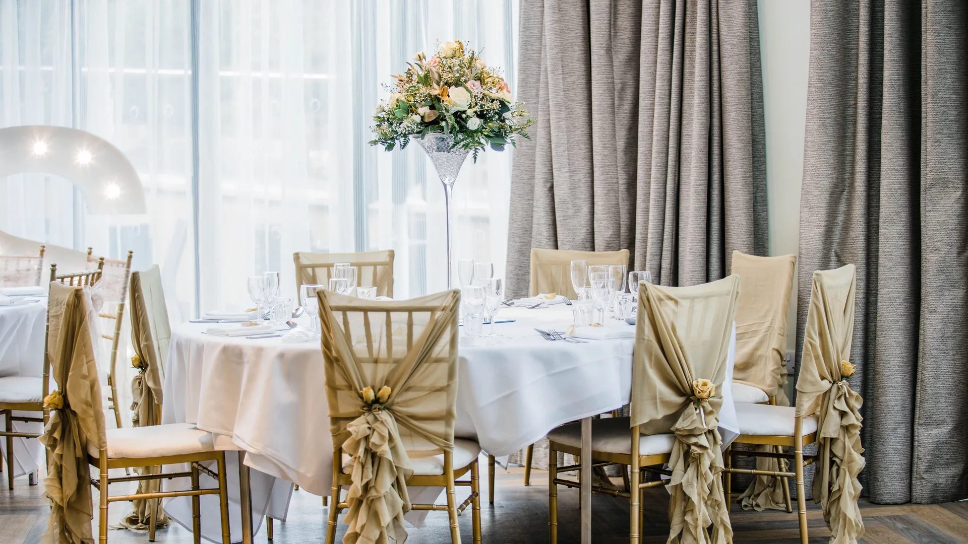 Elegant wedding reception table with gold Chiavari chairs, draped champagne covers and a tall floral centrepiece.