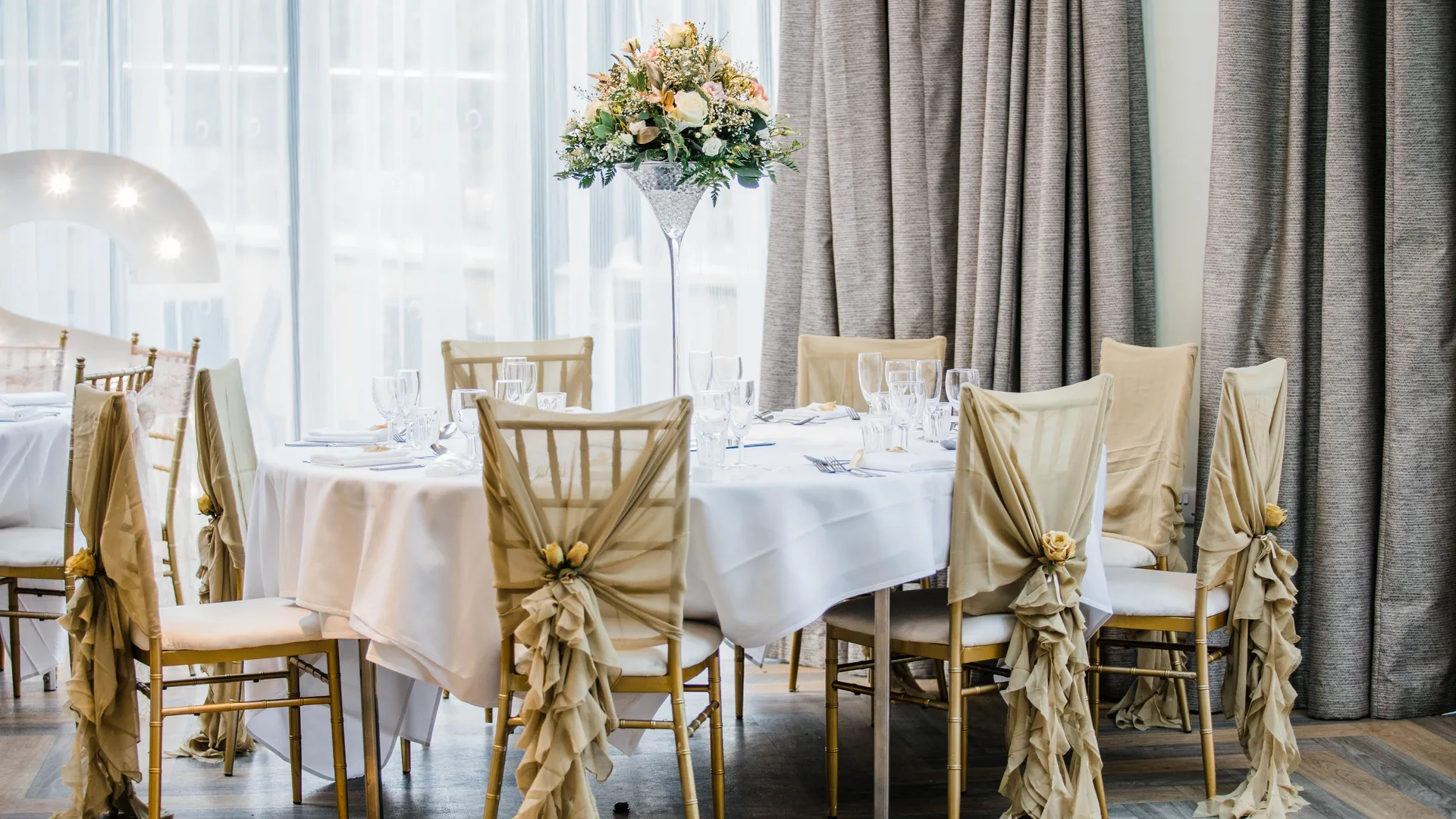 Elegant wedding reception table with gold Chiavari chairs, draped champagne covers and a tall floral centrepiece.