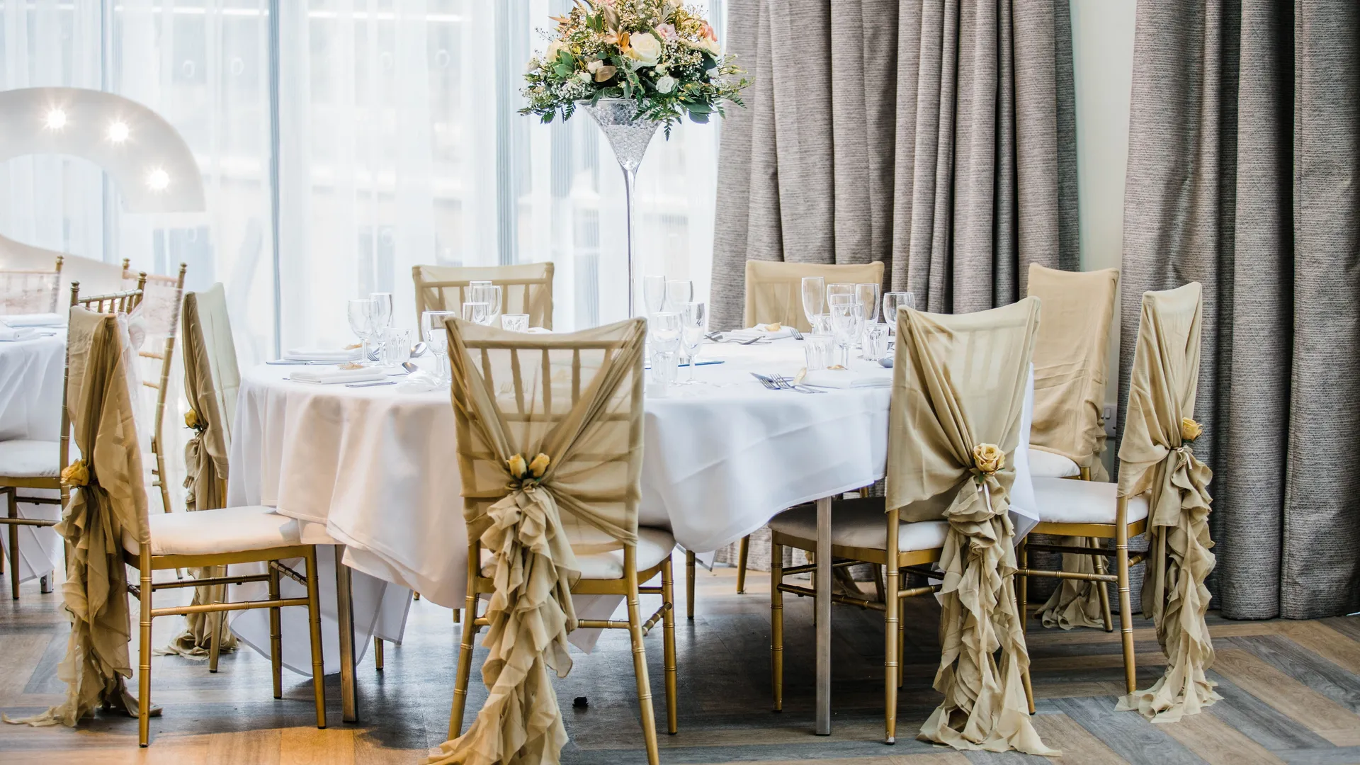 Elegant wedding reception table with gold Chiavari chairs, draped champagne covers and a tall floral centrepiece.