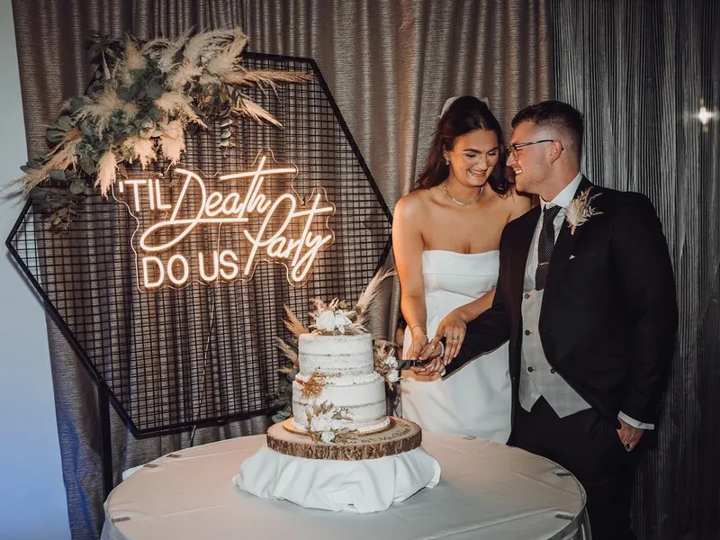 Bride and groom cutting a two-tier wedding cake beside a glowing 'Til Death Do Us Party' sign.