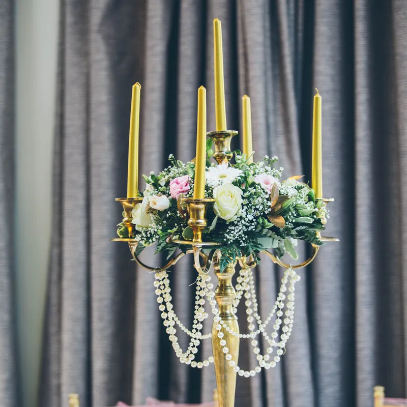 Elegant gold candelabra centrepiece with yellow taper candles, pastel flowers and pearl garlands on a formally set dining table.
