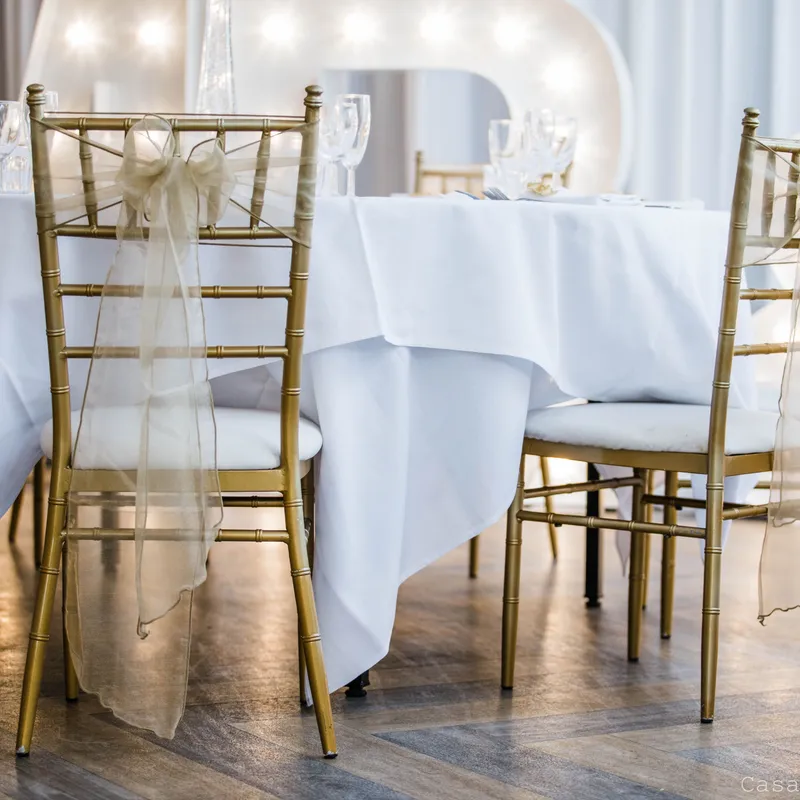 Gold Chiavari chairs with sheer ribbon bows beside a white-clothed wedding table and illuminated starlight letters.