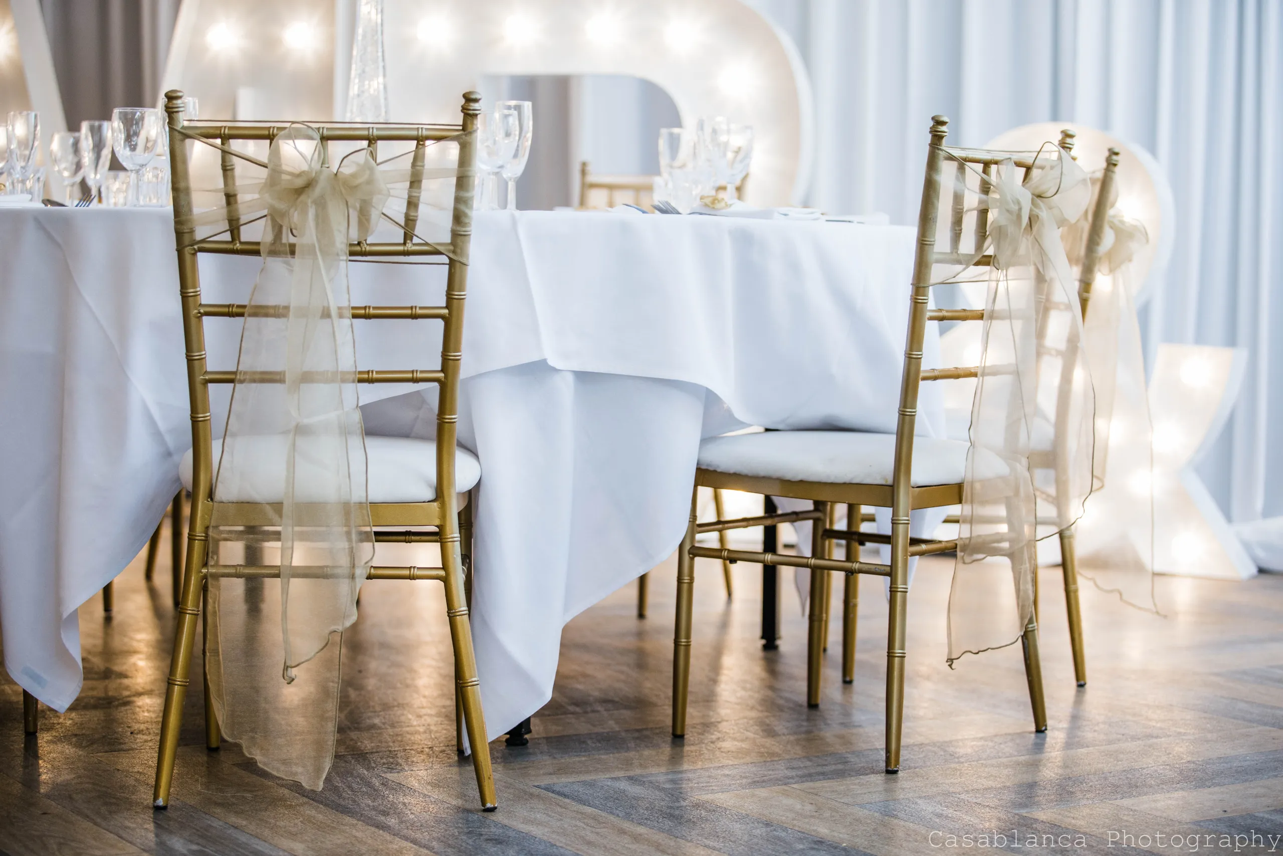 Gold Chiavari chairs with sheer ribbon bows beside a white-clothed wedding table and illuminated starlight letters.