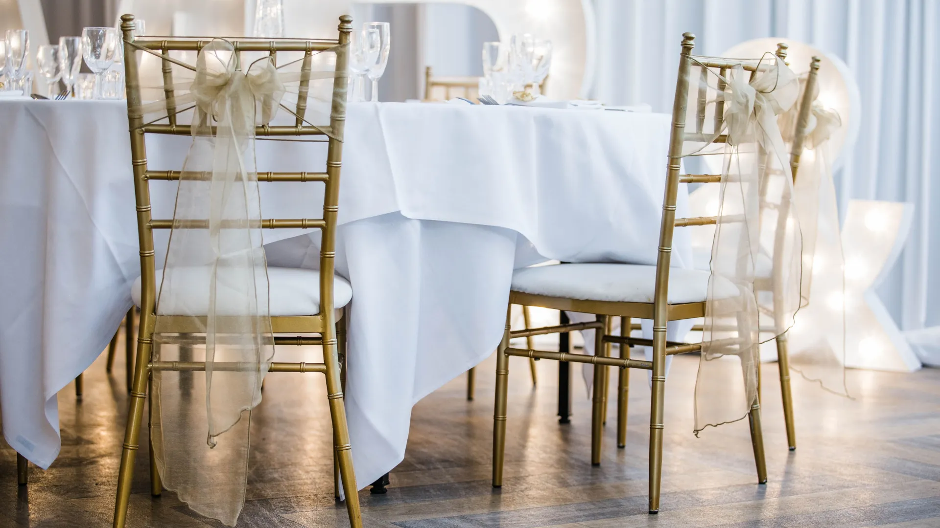 Gold Chiavari chairs with sheer ribbon bows beside a white-clothed wedding table and illuminated starlight letters.