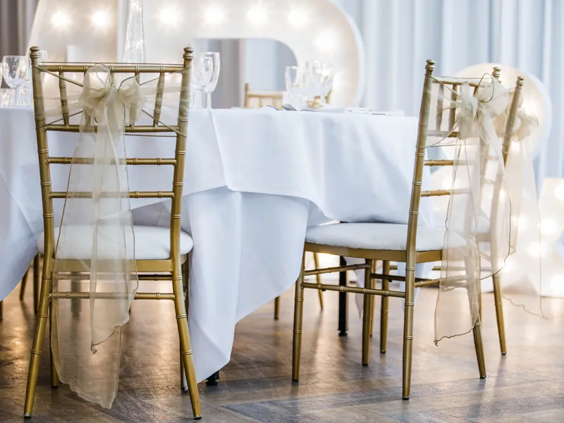 Gold Chiavari chairs with sheer ribbon bows beside a white-clothed wedding table and illuminated starlight letters.