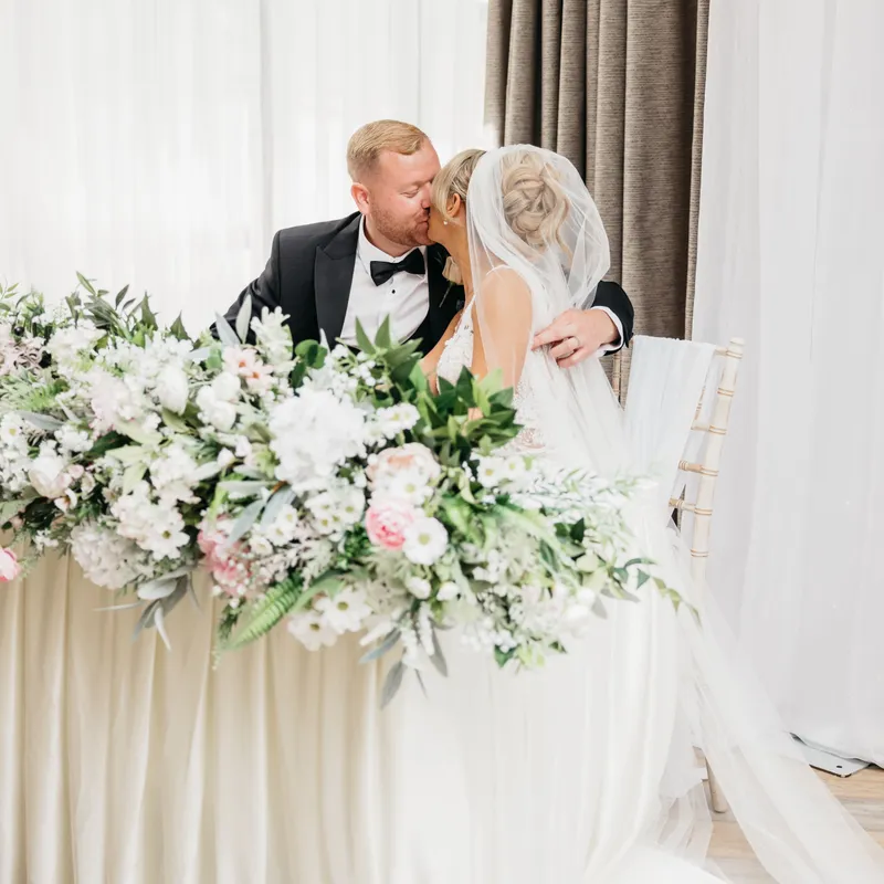 Newlywed couple sharing a kiss behind a flower-filled wedding table