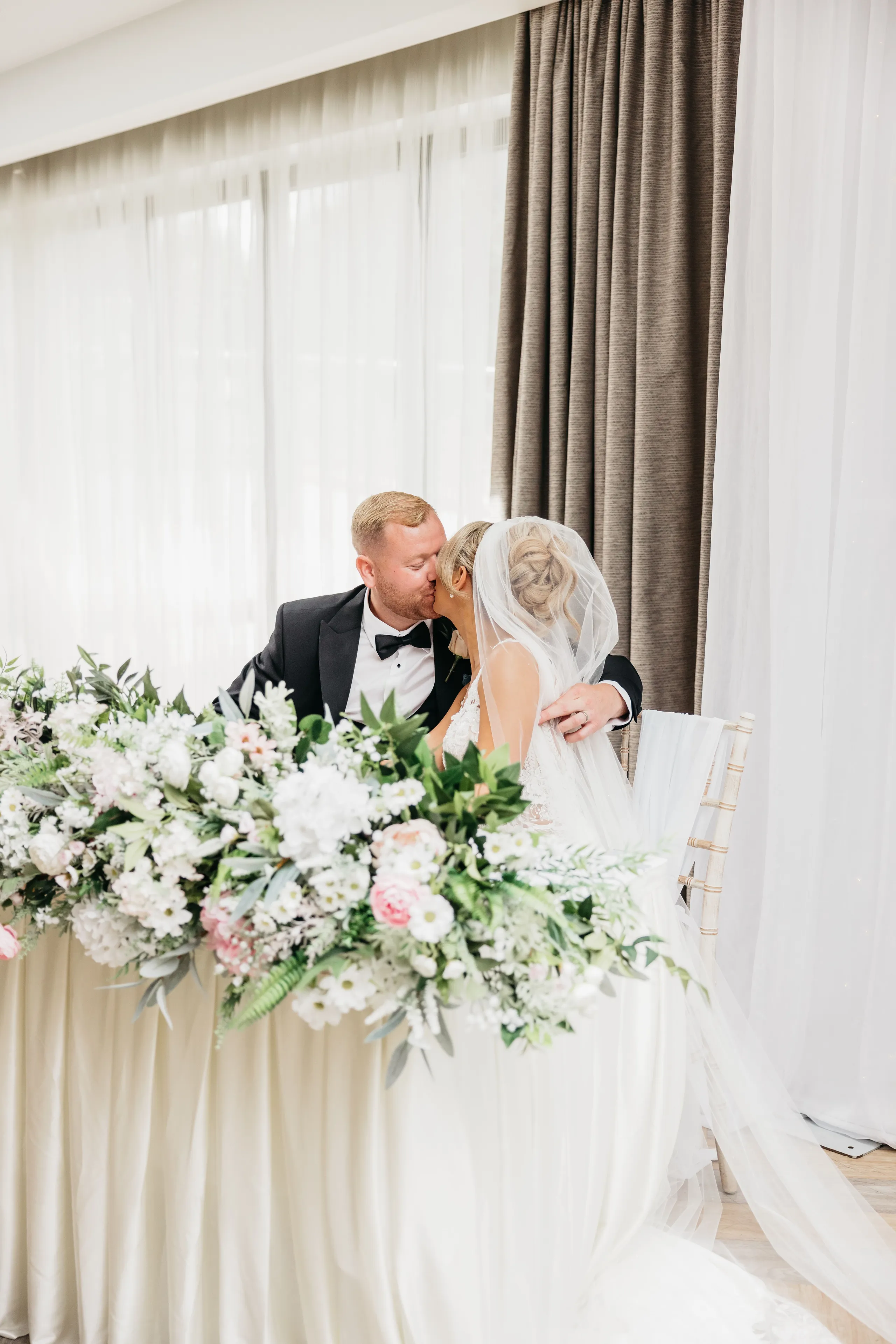 Newlywed couple sharing a kiss behind a flower-filled wedding table