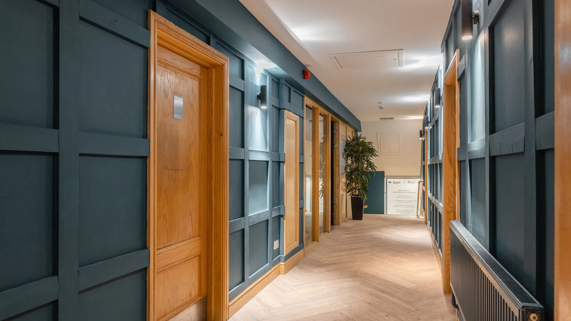 Modern interior corridor with dark blue panelled walls, wooden doors, wall lights and tiled flooring.