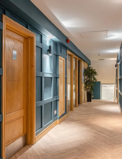 Modern interior corridor with dark blue panelled walls, wooden doors, wall lights and tiled flooring.