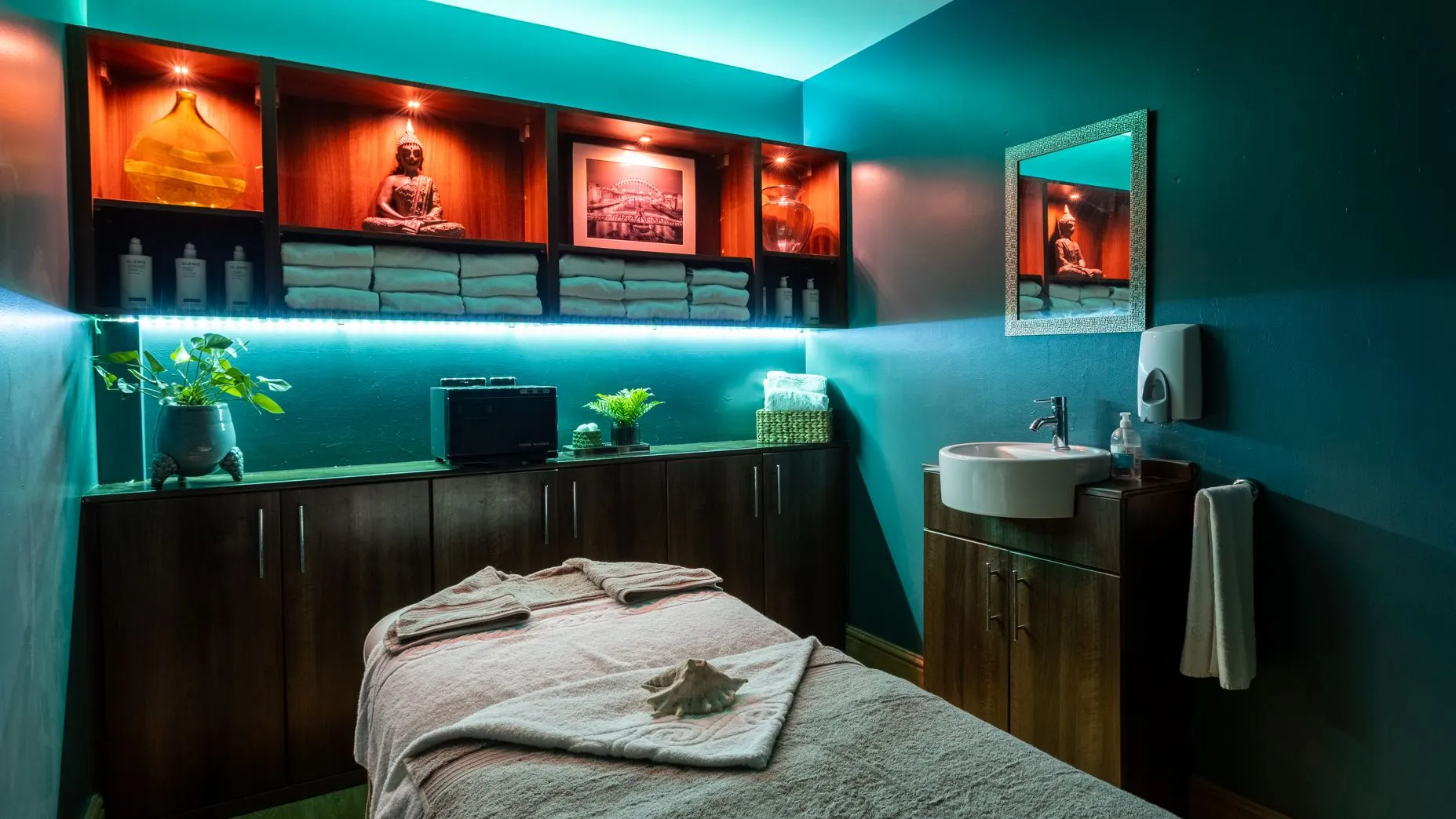 Tranquil spa treatment room with a massage bed, folded towels, Buddha statue and turquoise ambient lighting.