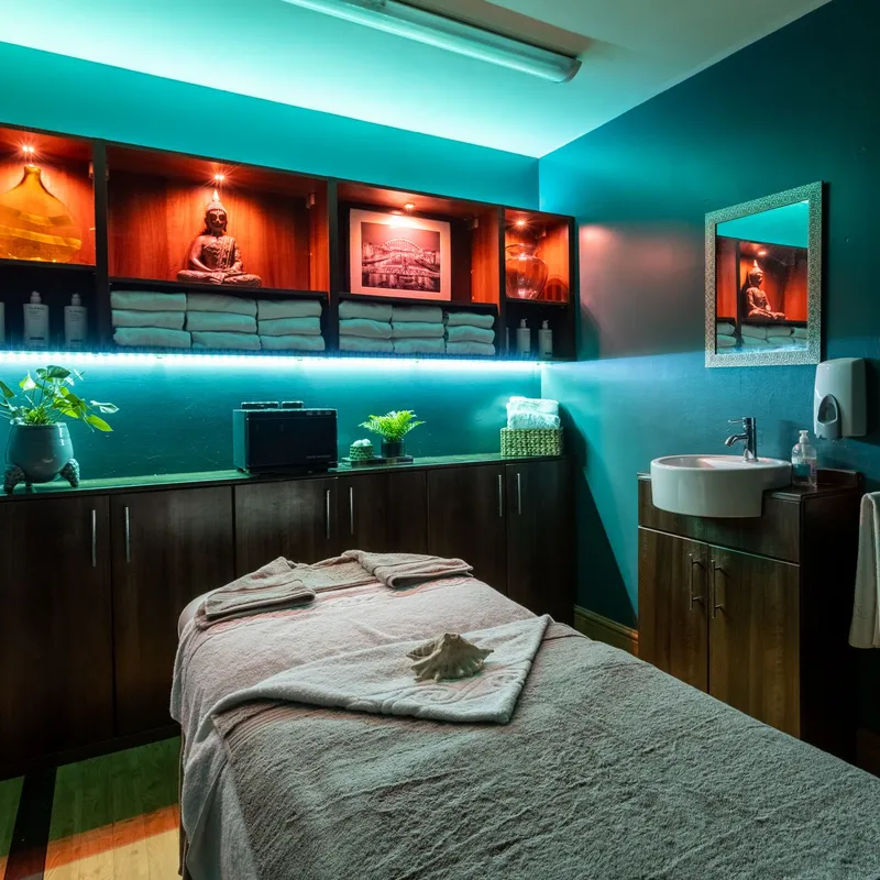 Tranquil spa treatment room with a massage bed, folded towels, Buddha statue and turquoise ambient lighting.