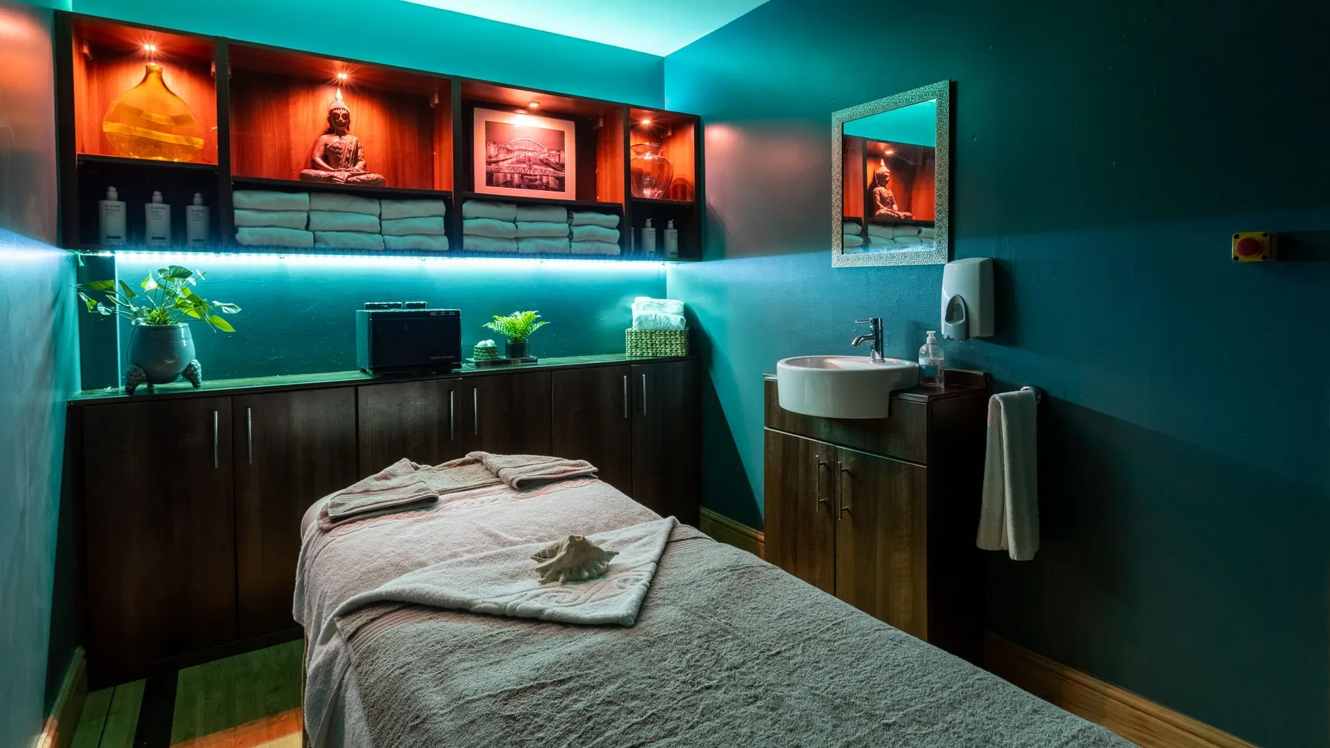 Tranquil spa treatment room with a massage bed, folded towels, Buddha statue and turquoise ambient lighting.