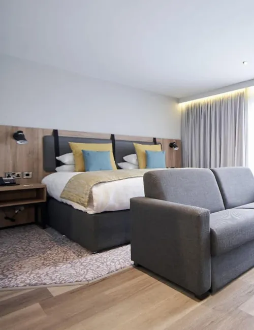 Modern hotel bedroom with a double bed, grey sofa and bright window seating area