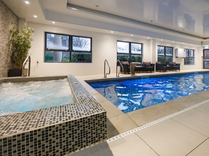 Modern indoor swimming pool with a bubbling tiled hot tub, lounge seating and large windows.