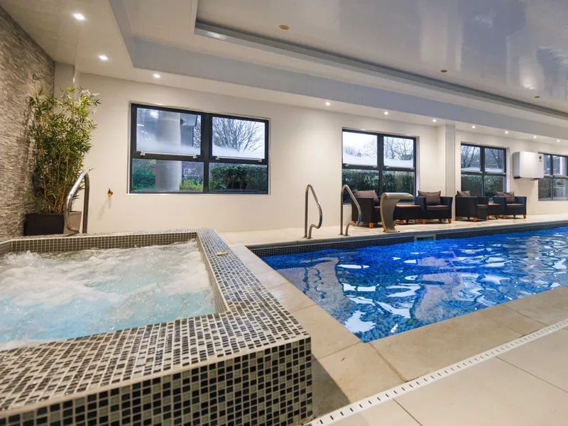 Modern indoor swimming pool with a bubbling tiled hot tub, lounge seating and large windows.