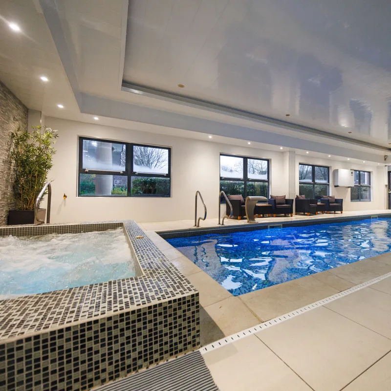 Modern indoor swimming pool with a bubbling tiled hot tub, lounge seating and large windows.