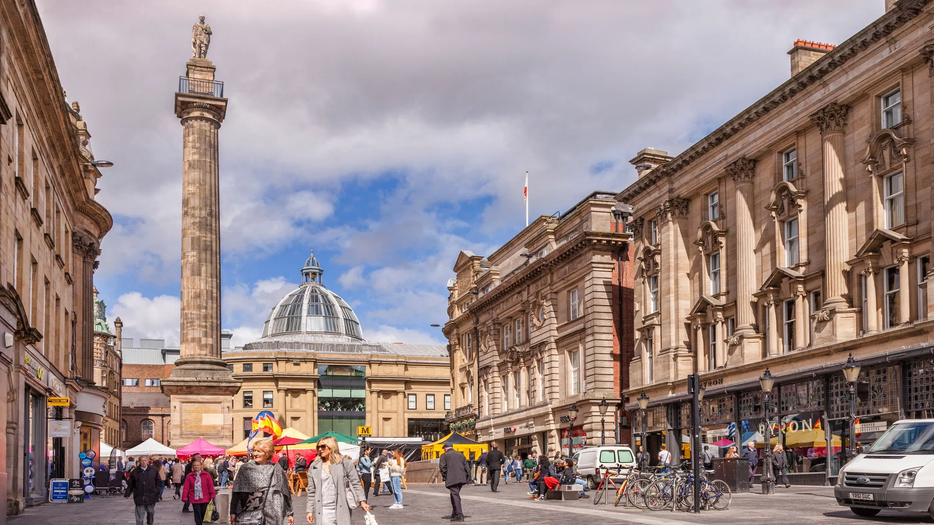 Pedestrians walk through a busy square in Birmingham city centre beside a tall monument column and grand historic buildings.