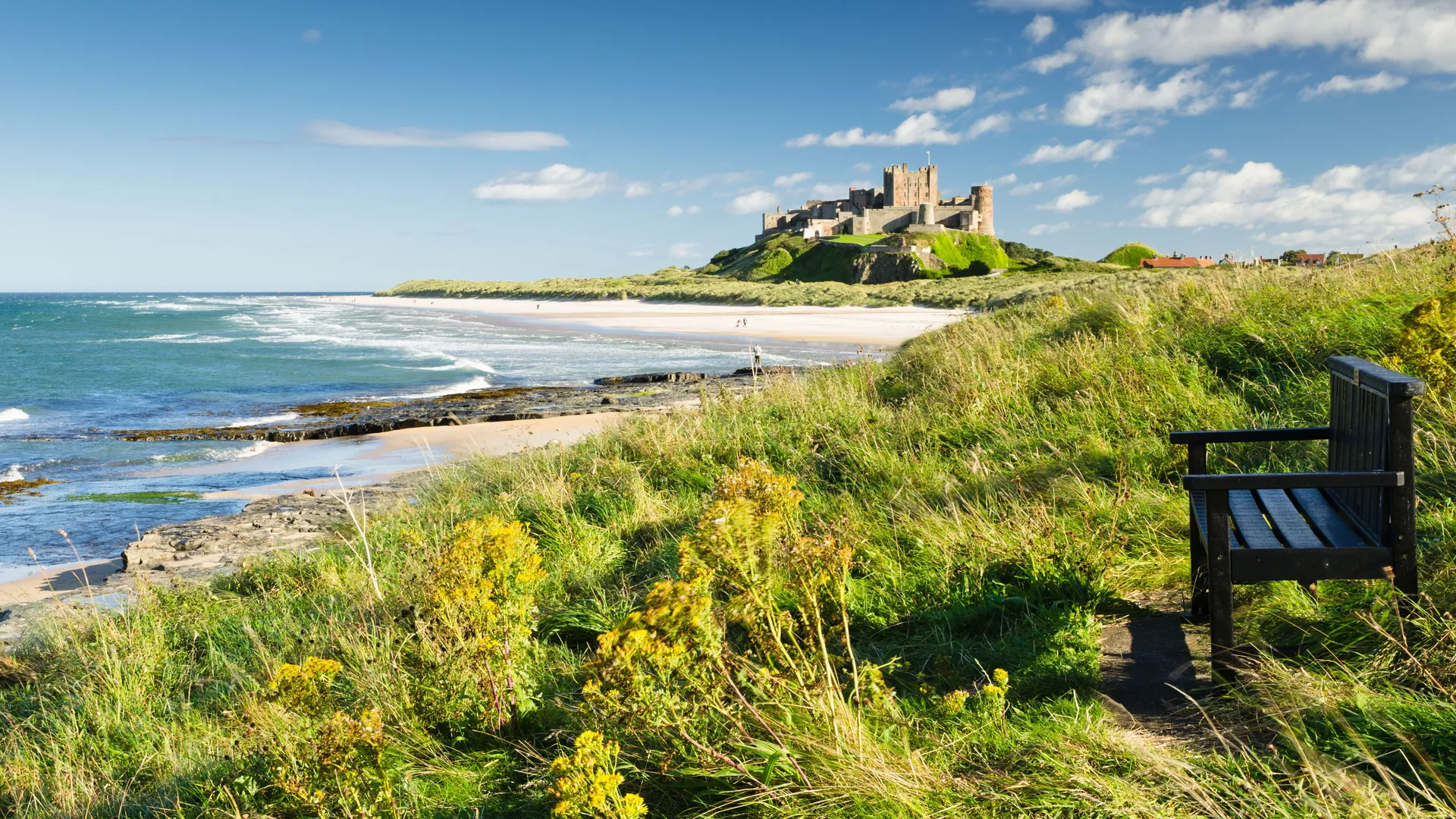Wooden bench overlooking Bamburgh Castle, sandy beach and the North Sea under a blue sky.