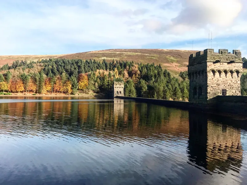 Stone dam tower reflected in a calm reservoir, with autumn woodland and hills behind
