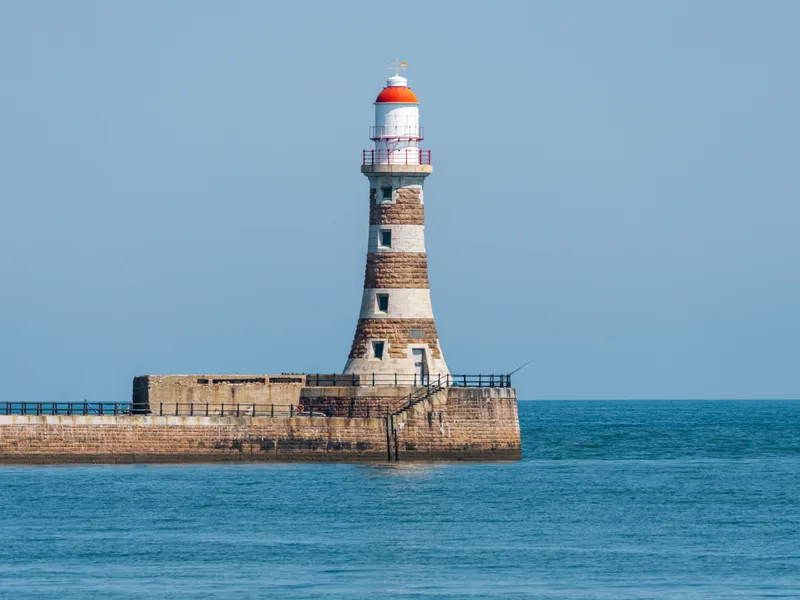 Stone lighthouse with a red lantern roof standing on a sea wall beneath a clear blue sky