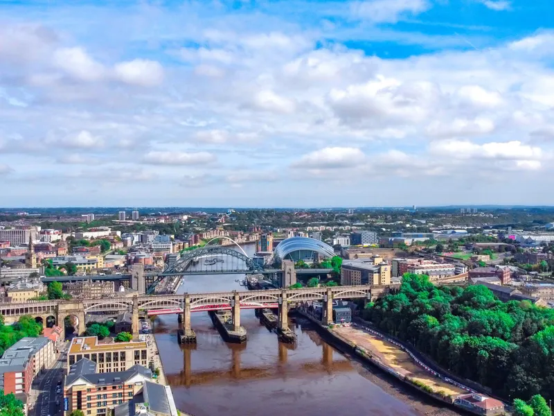 Aerial view of Newcastle upon Tyne with the River Tyne and its bridges under a cloudy blue sky.