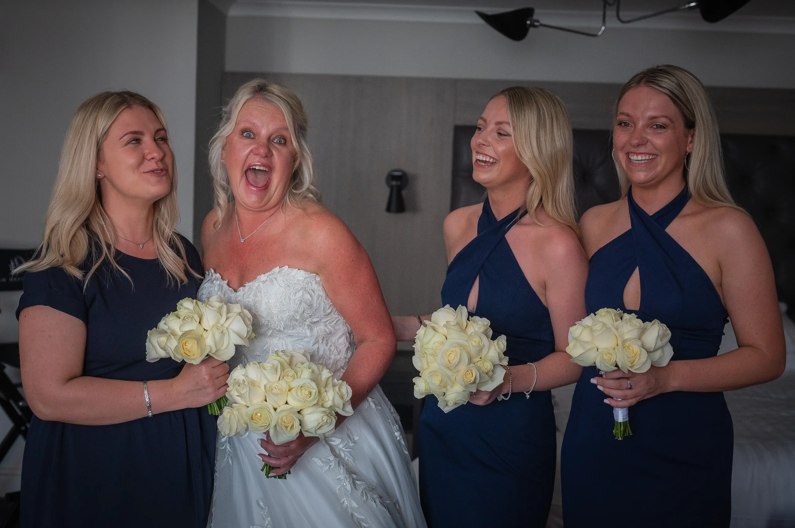 Bride laughing with three bridesmaids holding cream rose bouquets in a hotel room.