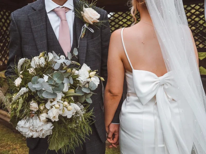 Bride in a white bow-backed wedding dress and veil holding hands with groom in a dark checked suit, carrying a white floral bouquet.