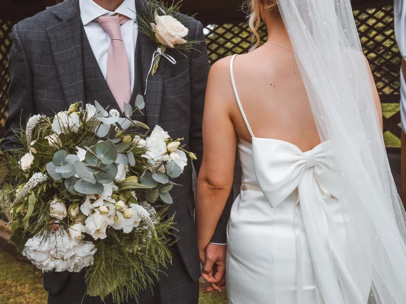 Bride in a white bow-backed wedding dress and veil holding hands with groom in a dark checked suit, carrying a white floral bouquet.