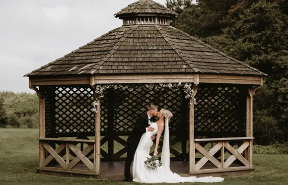 Bride and groom kissing beneath a rustic wooden gazebo in a grassy garden