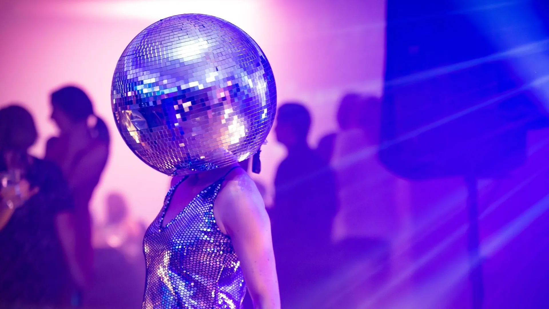 Person wearing a mirrored disco-ball helmet and shimmering dress at a purple-lit party