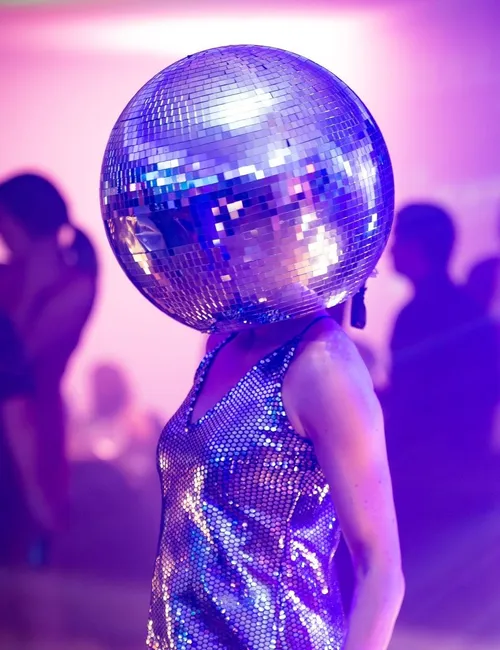 Person wearing a mirrored disco-ball helmet and shimmering dress at a purple-lit party