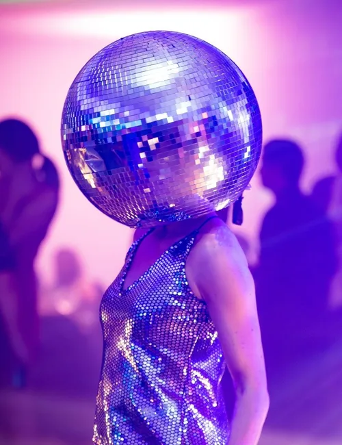 Person wearing a mirrored disco-ball helmet and shimmering dress at a purple-lit party