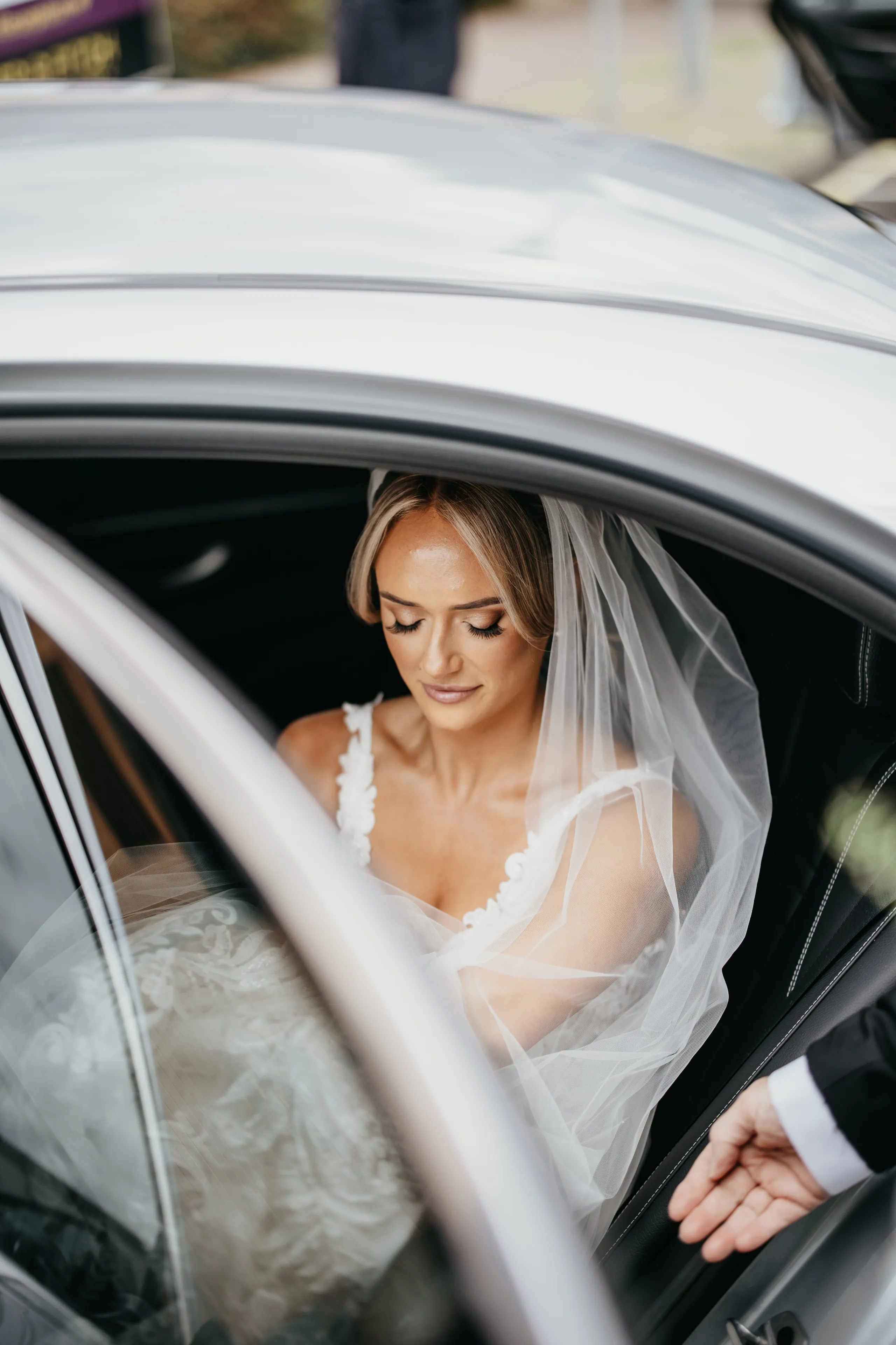 Bride in a lace wedding dress and veil seated in a car, with a hand opening the door.