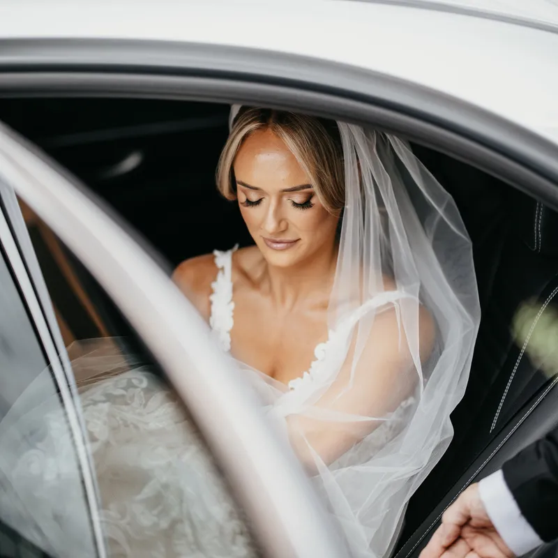 Bride in a lace wedding dress and veil seated in a car, with a hand opening the door.