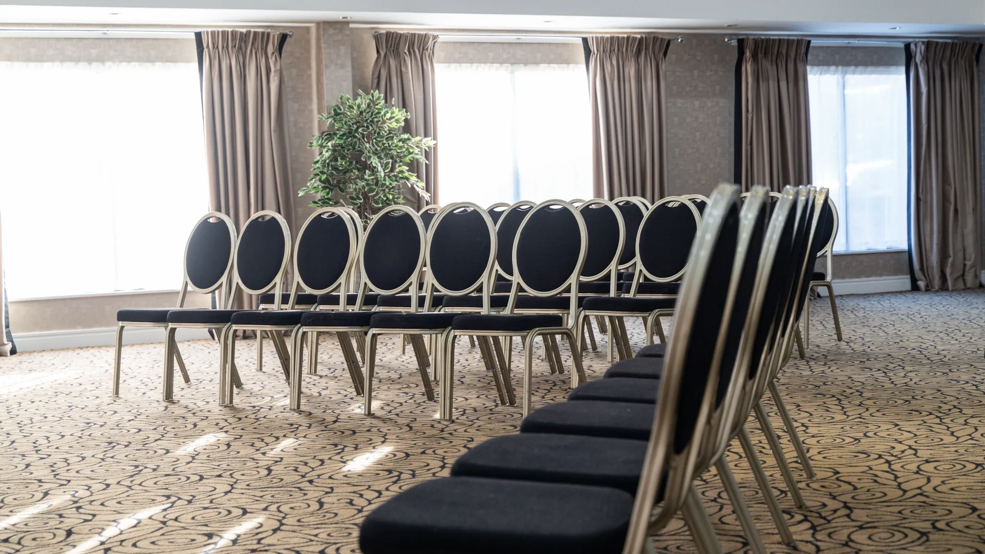Rows of black upholstered chairs arranged in a bright hotel meeting room with large curtained windows.