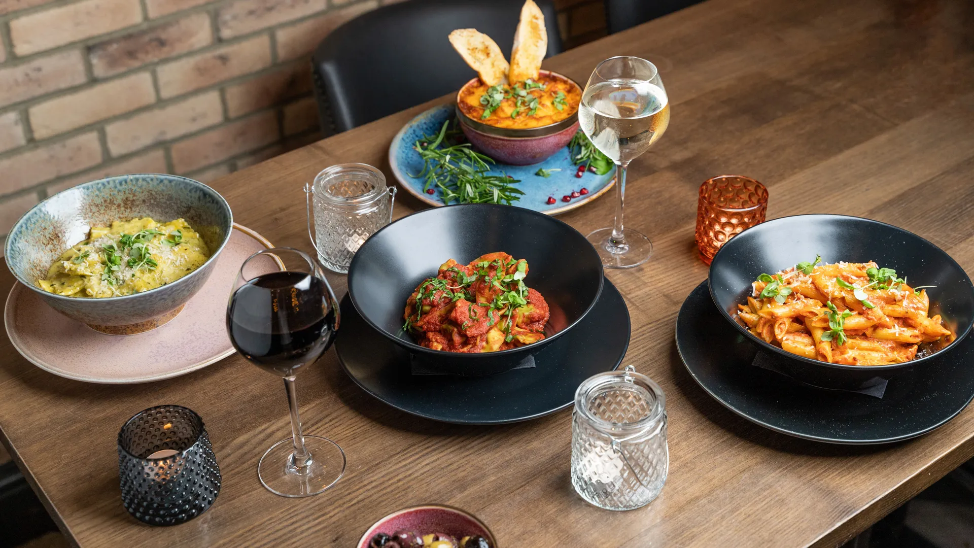 Wooden restaurant table set with colourful pasta dishes, wine glasses and candle holders.