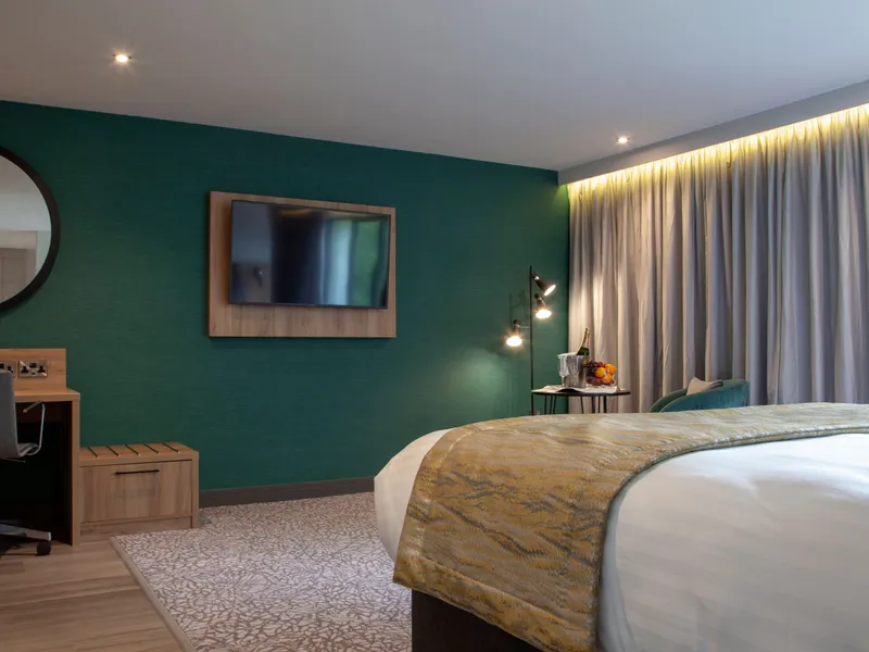 Modern hotel bedroom with teal feature wall, king-size bed, desk and wall-mounted television.