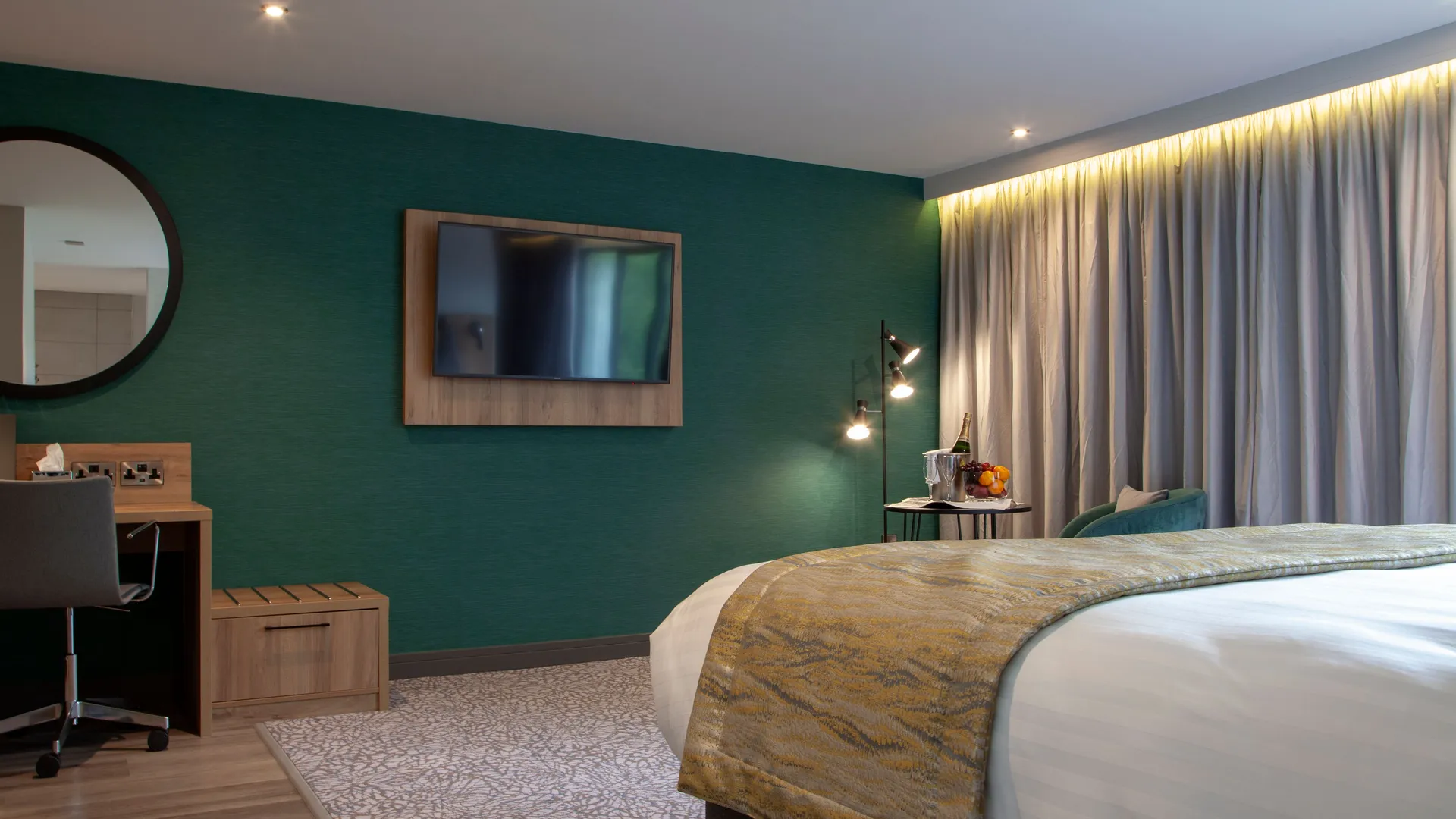 Modern hotel bedroom with teal feature wall, king-size bed, desk and wall-mounted television.
